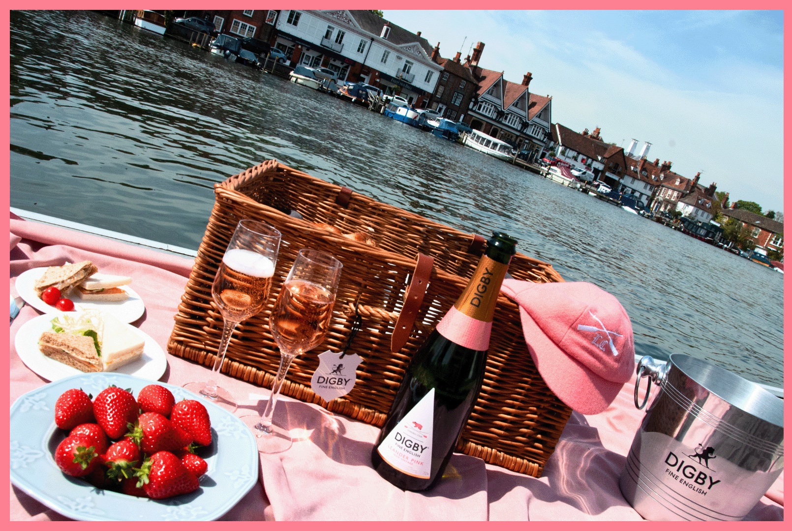 A bottle of Digby's Leander Pink on a picnic rug with strawberries and a Leander Club cap, on the banks of the River Thames in Henley.